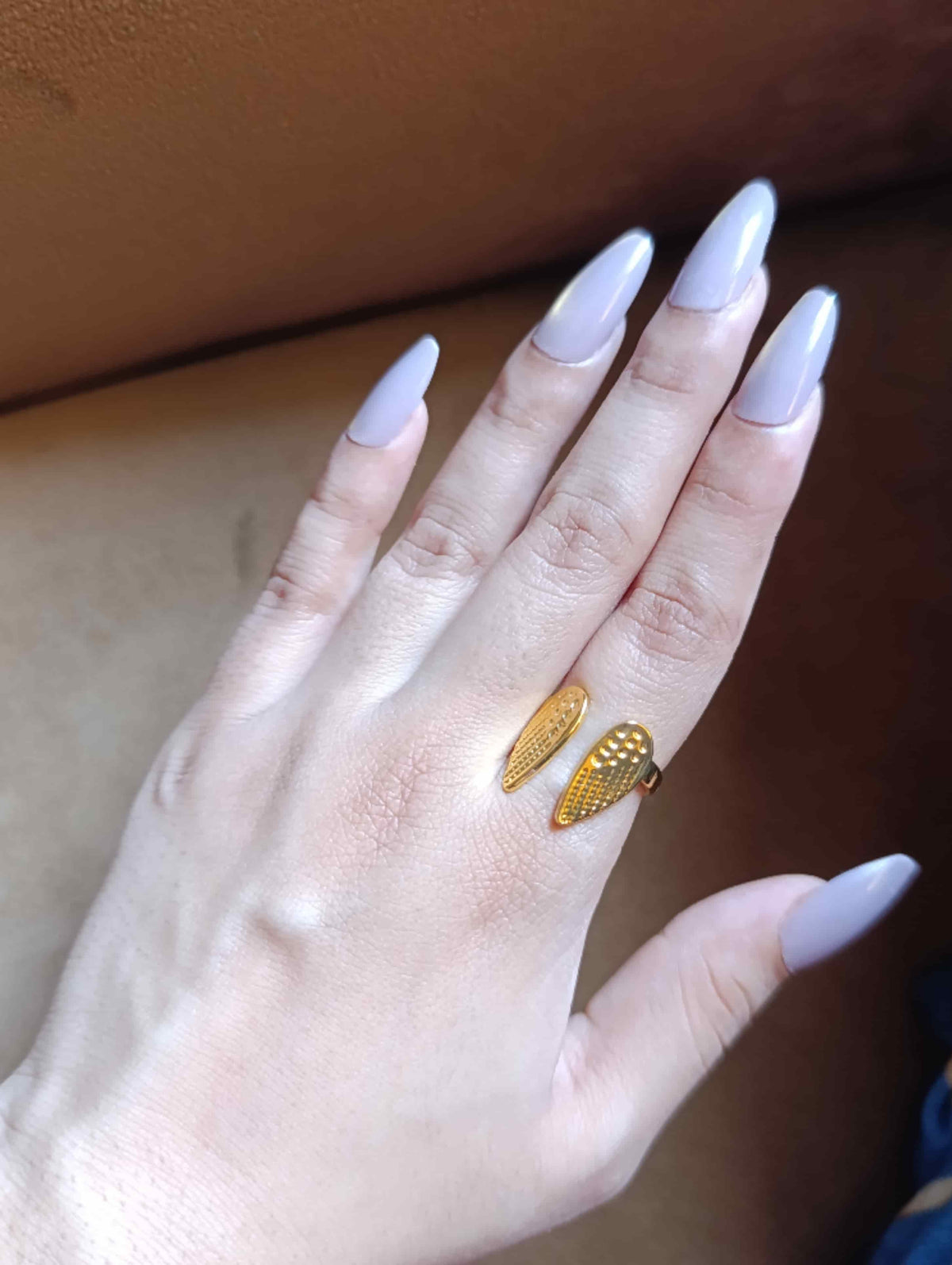 A close-up of a hand with long, light lavender-painted nails wearing a gold ring shaped like two textured wings on the ring finger, set against a brown leather background.