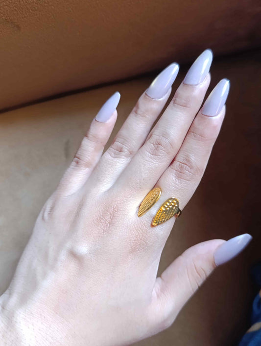 A close-up of a hand with long, light lavender-painted nails wearing a gold ring shaped like two textured wings on the ring finger, set against a brown leather background.