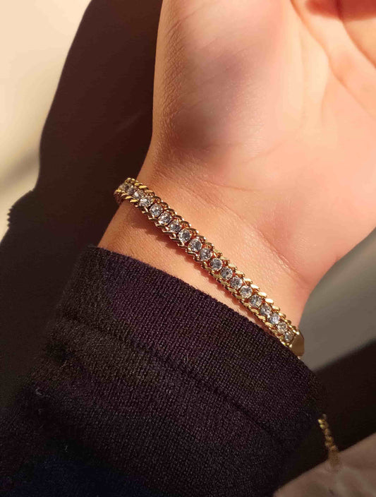 A gold tennis bracelet with clear stones worn on a person's wrist, shown against a dark sleeve and neutral background.