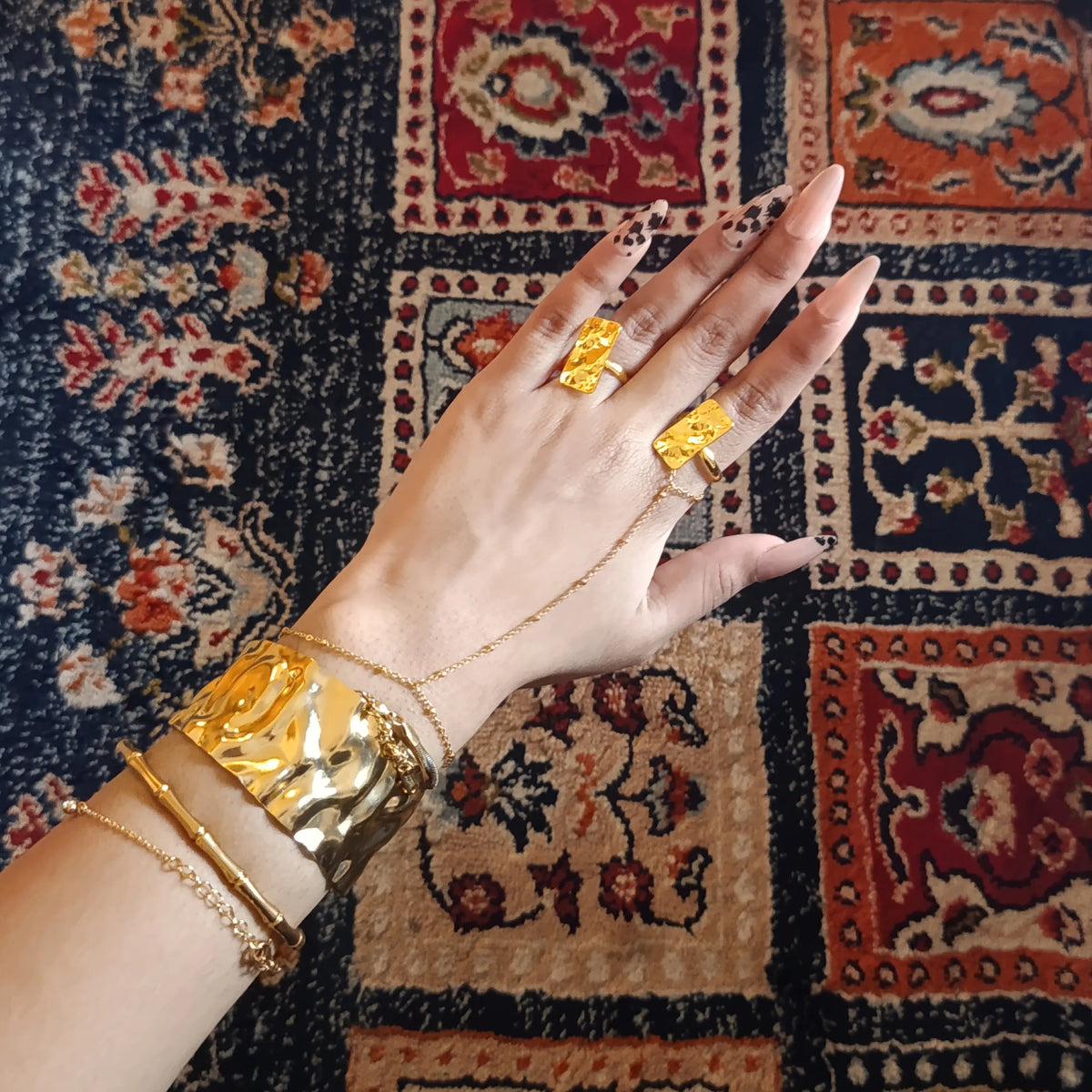 A hand wearing multiple gold jewelry pieces, including a large ring, a chain bracelet, and a chunky cuff bracelet, resting over a patterned rug.