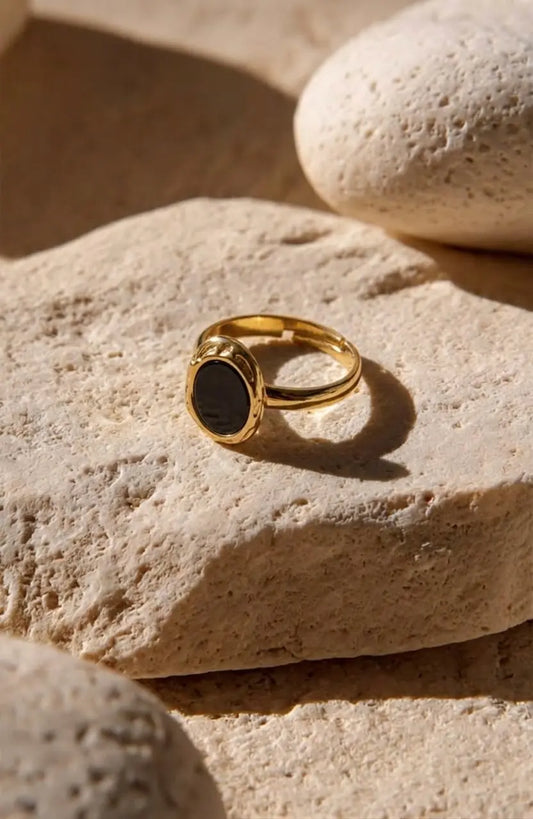 A gold signet ring with a dark oval stone, resting on a textured beige stone with other stones in the background.