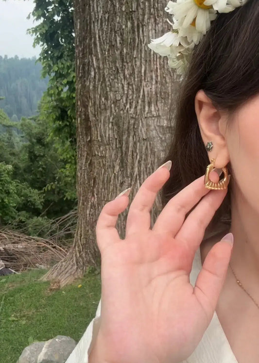 A close-up lifestyle shot of a person outdoors wearing a small gold heart-shaped hoop earring with a clear crystal center. The person is wearing a white top and a white daisy flower crown, with a large tree trunk and green forest in the background.