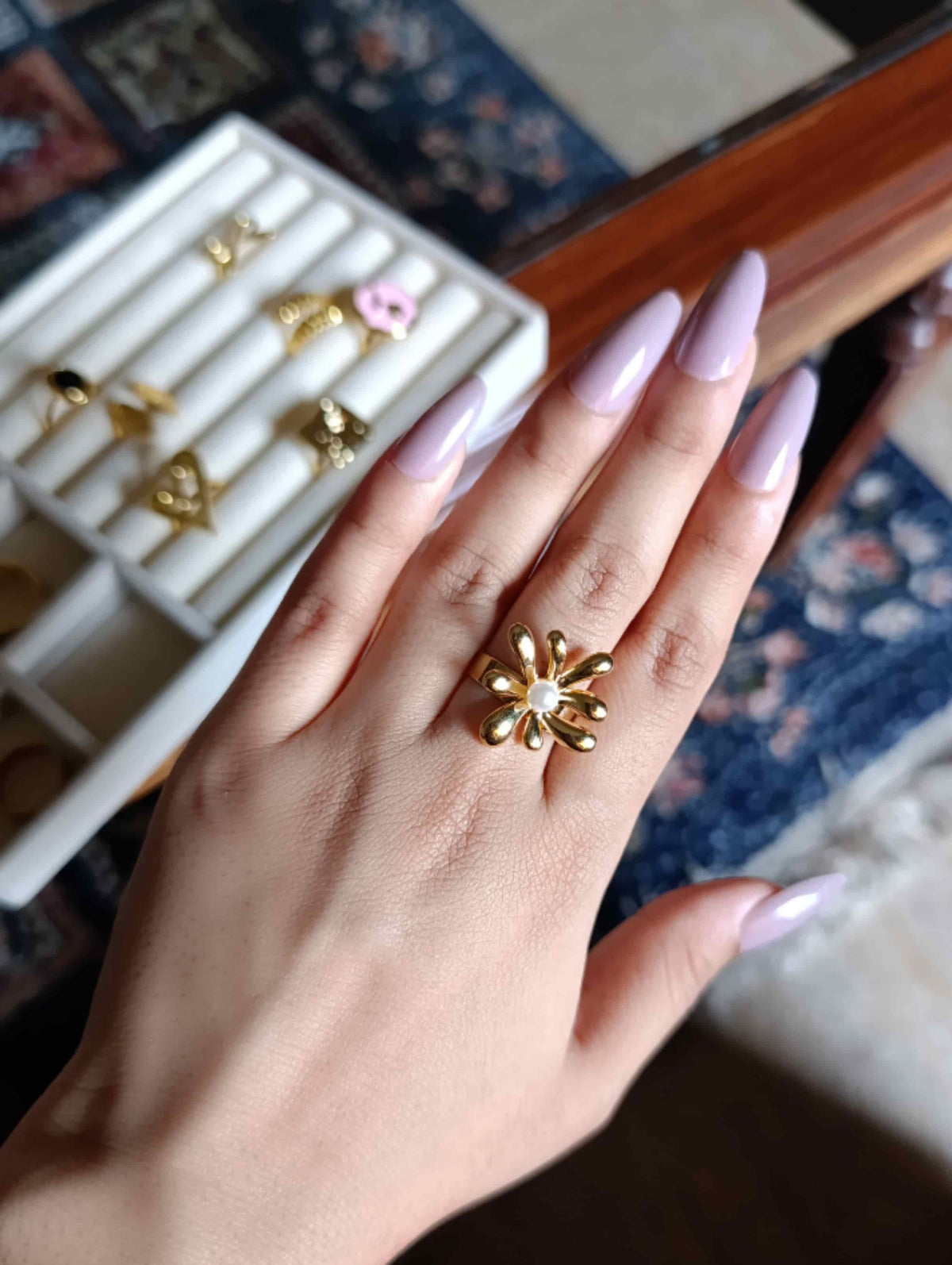 A close-up of a hand with lavender-painted nails wearing a gold flower-shaped ring, with a white jewelry organizer box in the background.