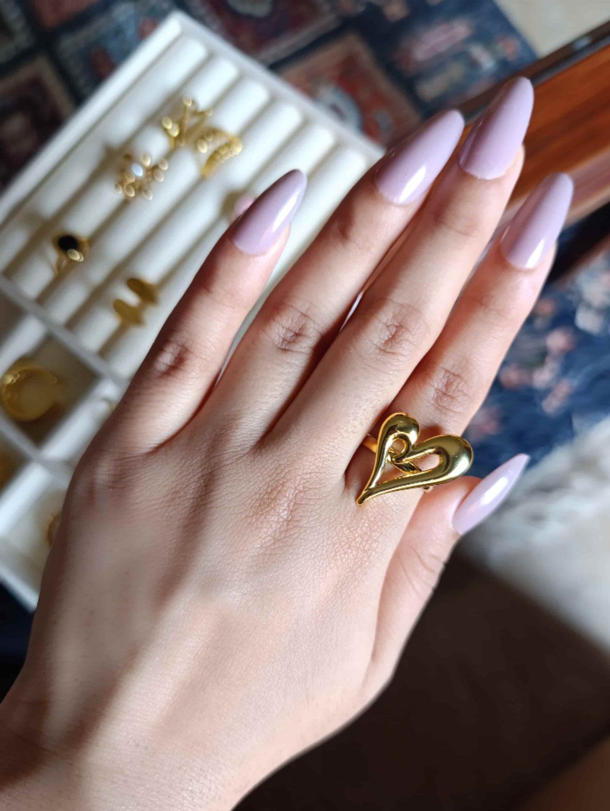 A close-up of a hand with lavender-painted nails wearing a gold heart-shaped ring, with a ring display tray visible in the background.