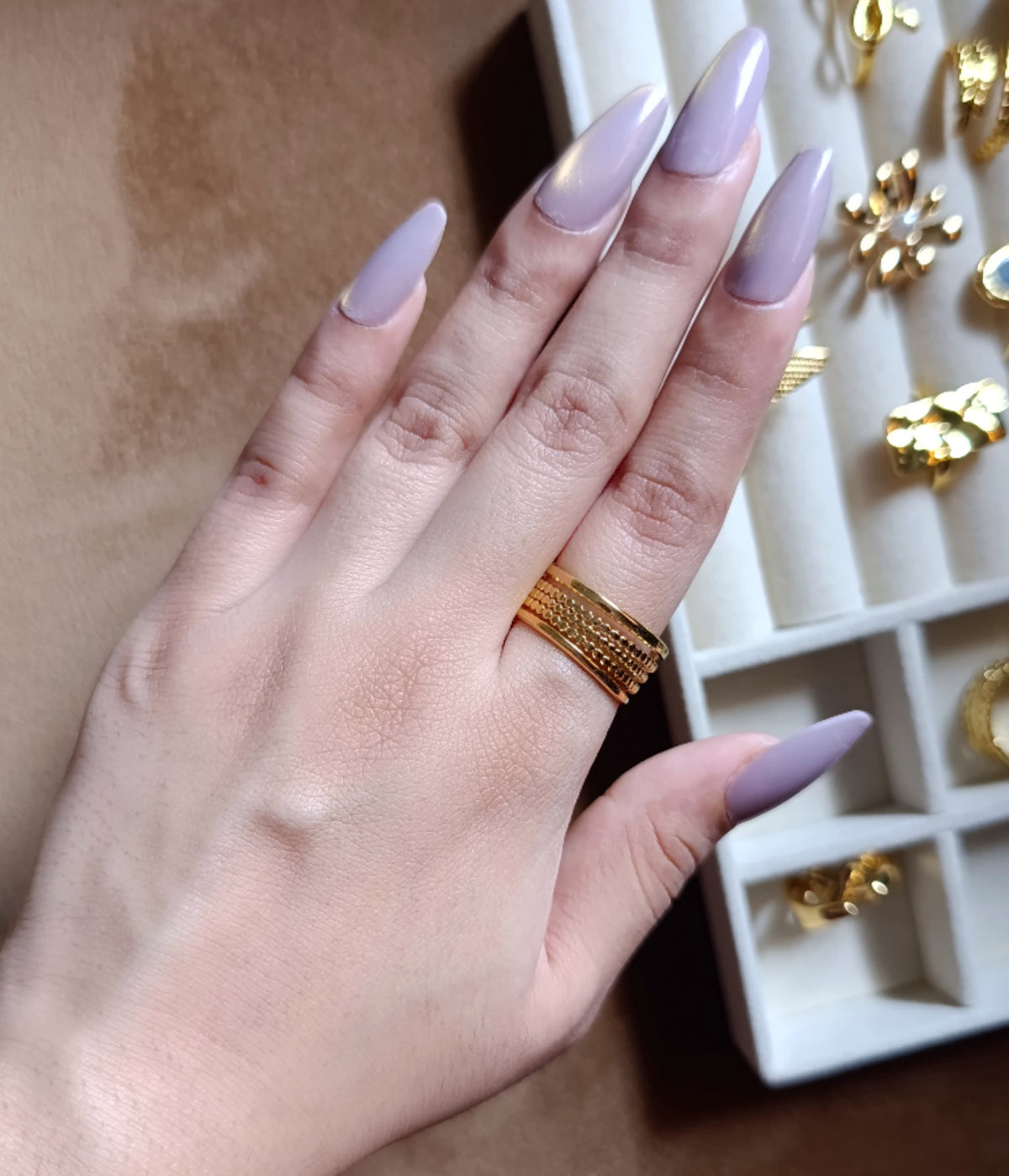 A close-up of a hand with lavender manicured nails wearing a wide textured gold ring, with a white ring display box containing other gold rings in the background.