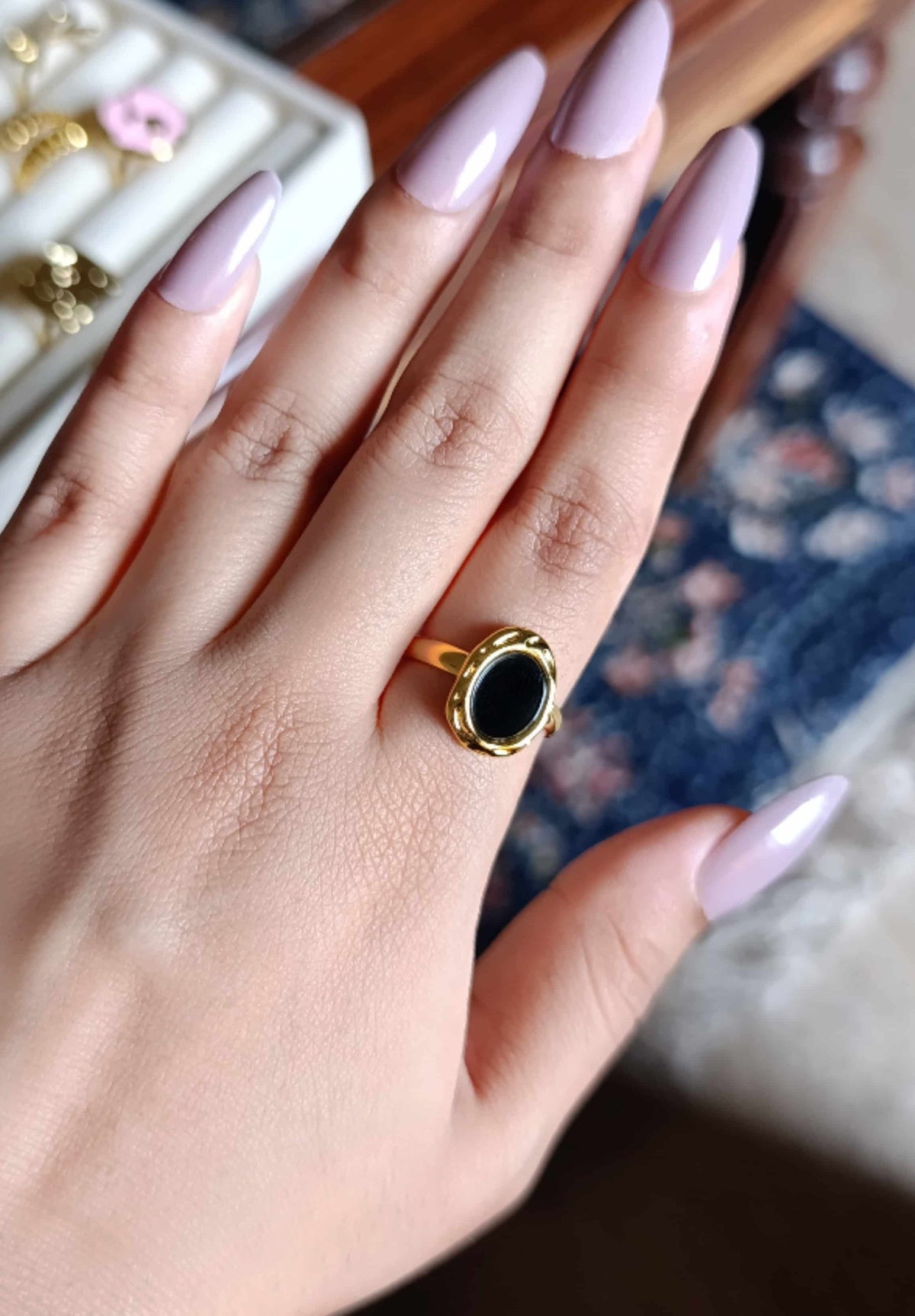 A close-up of a hand with long lavender manicured nails wearing a gold ring with a black oval stone. In the blurred background, a white jewelry tray with other rings is visible on a wooden table.