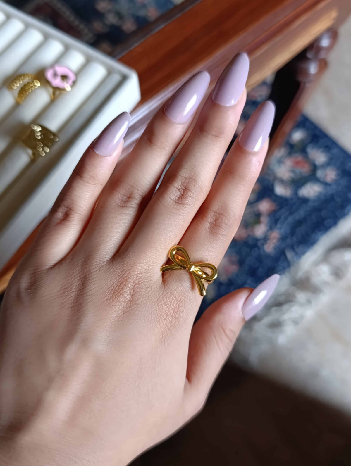 A close-up of a hand with long, light lavender manicured nails, wearing a shiny gold bow-shaped ring on the ring finger. The background shows a white jewelry display box with other rings and a patterned blue rug.