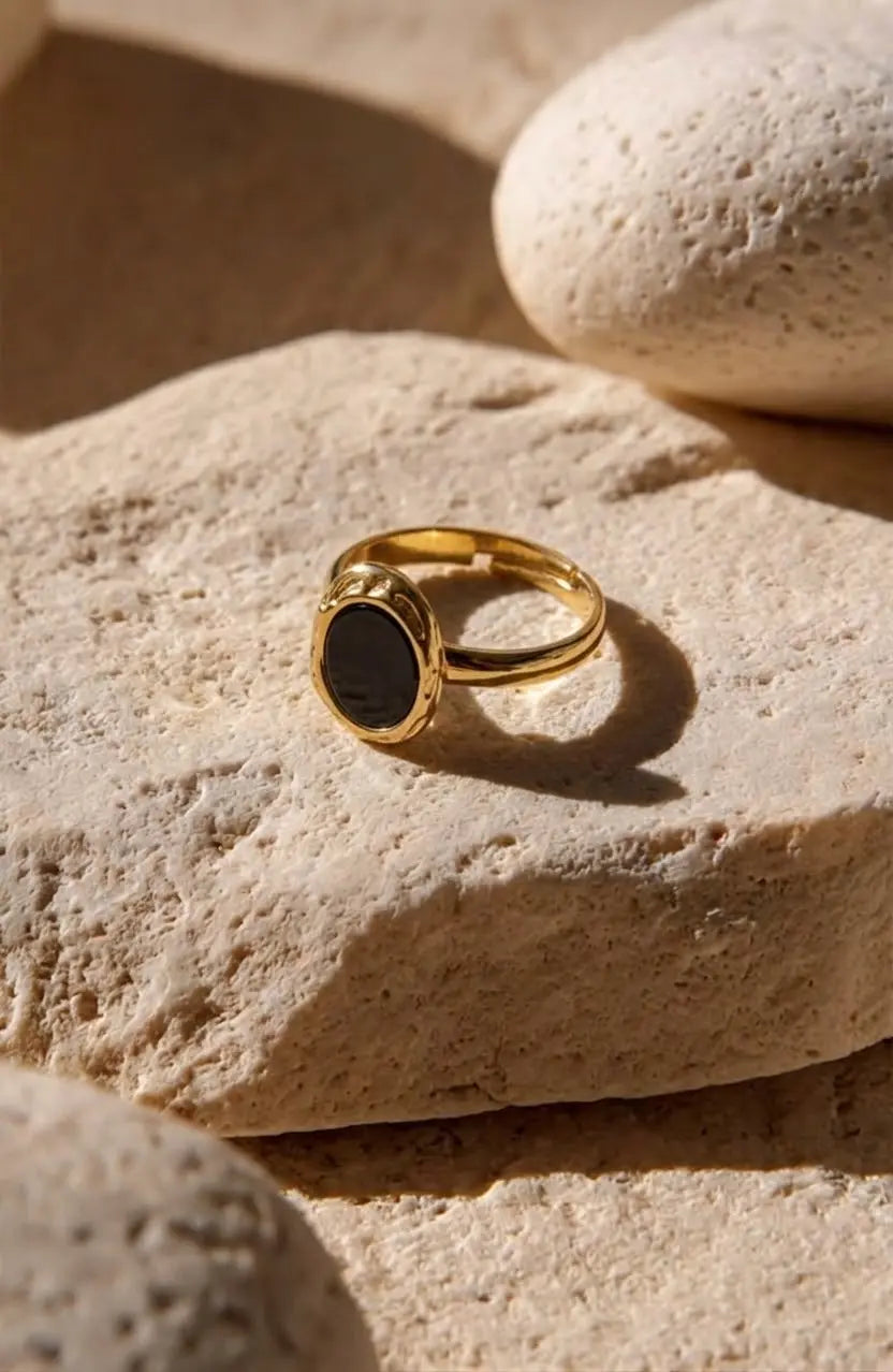 A gold signet ring with a dark oval stone, resting on a textured beige stone with other stones in the background.