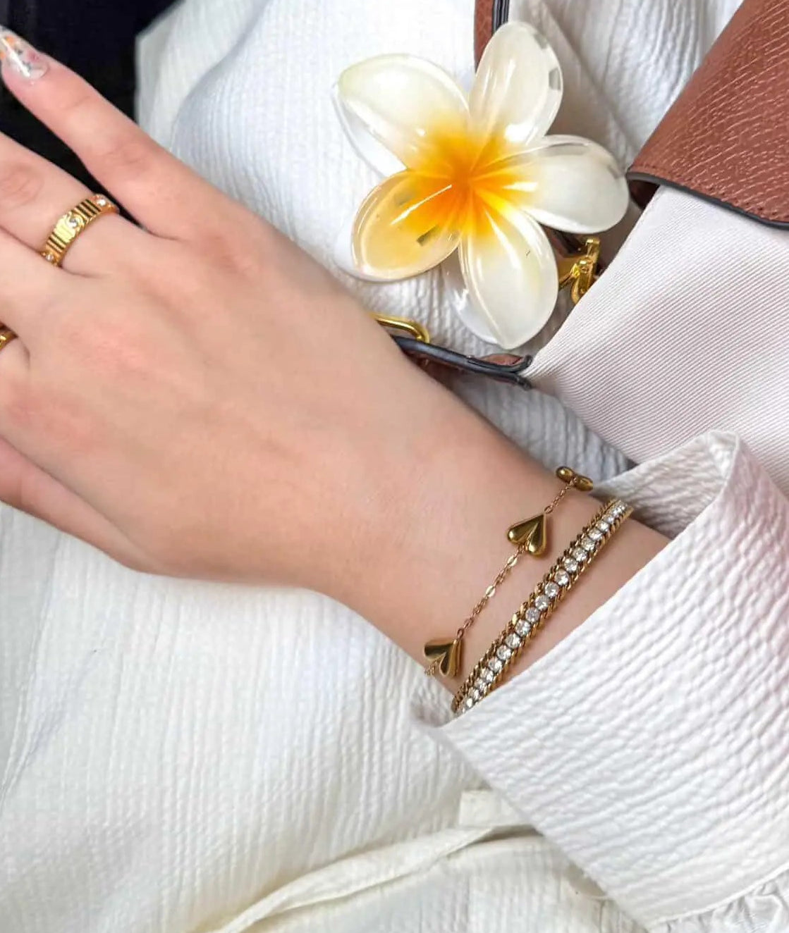 A close-up lifestyle shot of a person's hand and wrist wearing gold jewelry, including a gold ring, stacked gold bracelets, and a gold heart charm bracelet, styled with a white textured blouse and a white plumeria flower accessory.