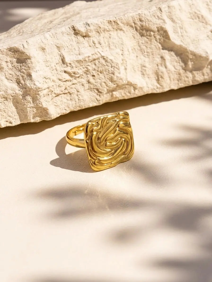 A gold ring with a highly textured, swirling, organic patterned face, displayed on a light beige surface with a large natural stone in the background.