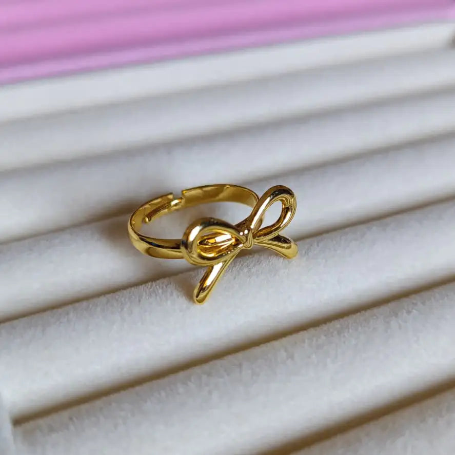 Gold bow-shaped ring on a white jewelry display with a pink background - Coquette Ring - Glitters For Her