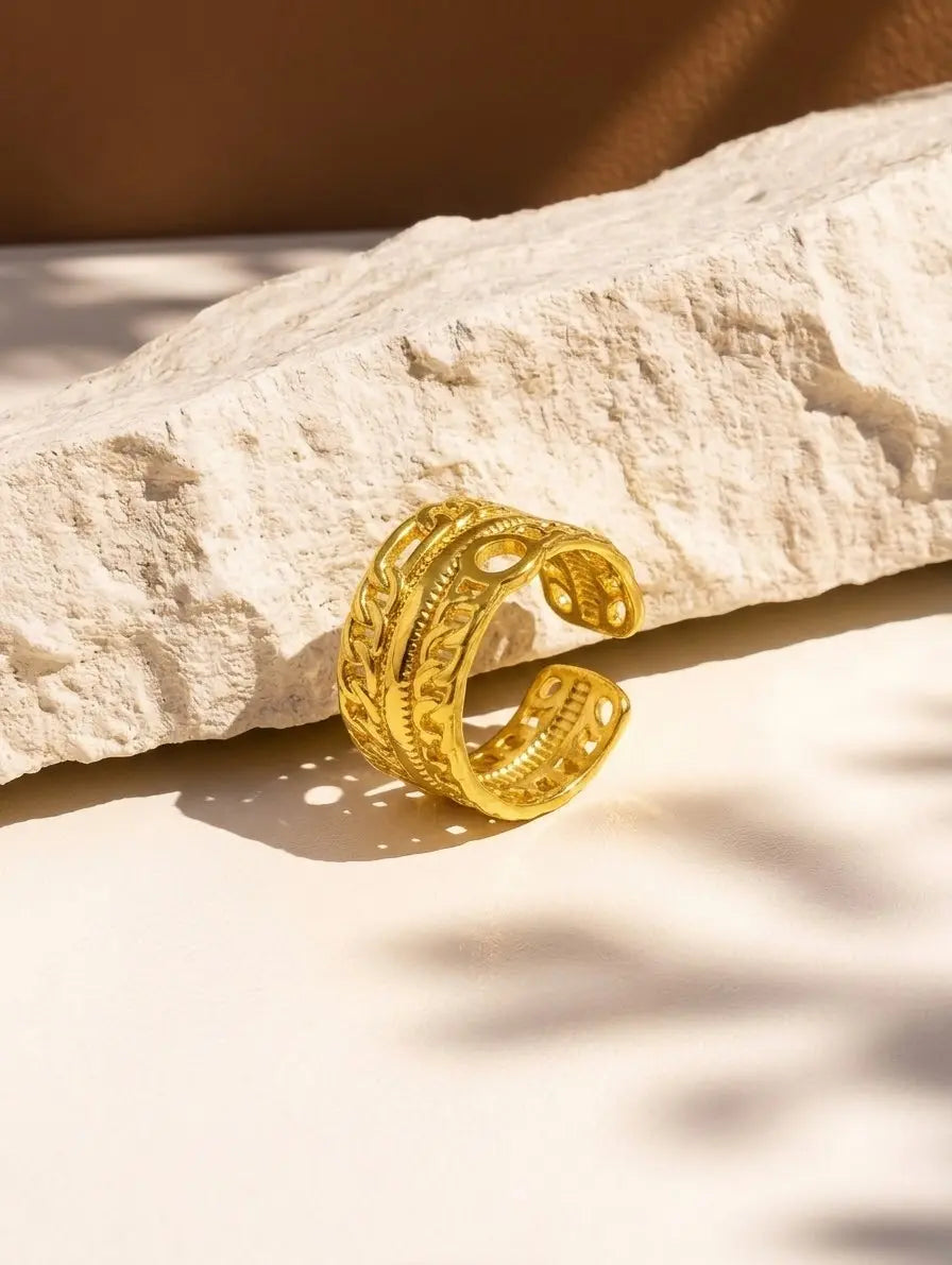 A gold-toned open cuff ring with intricate filigree cutouts, resting on a textured beige stone slab with warm natural shadows.